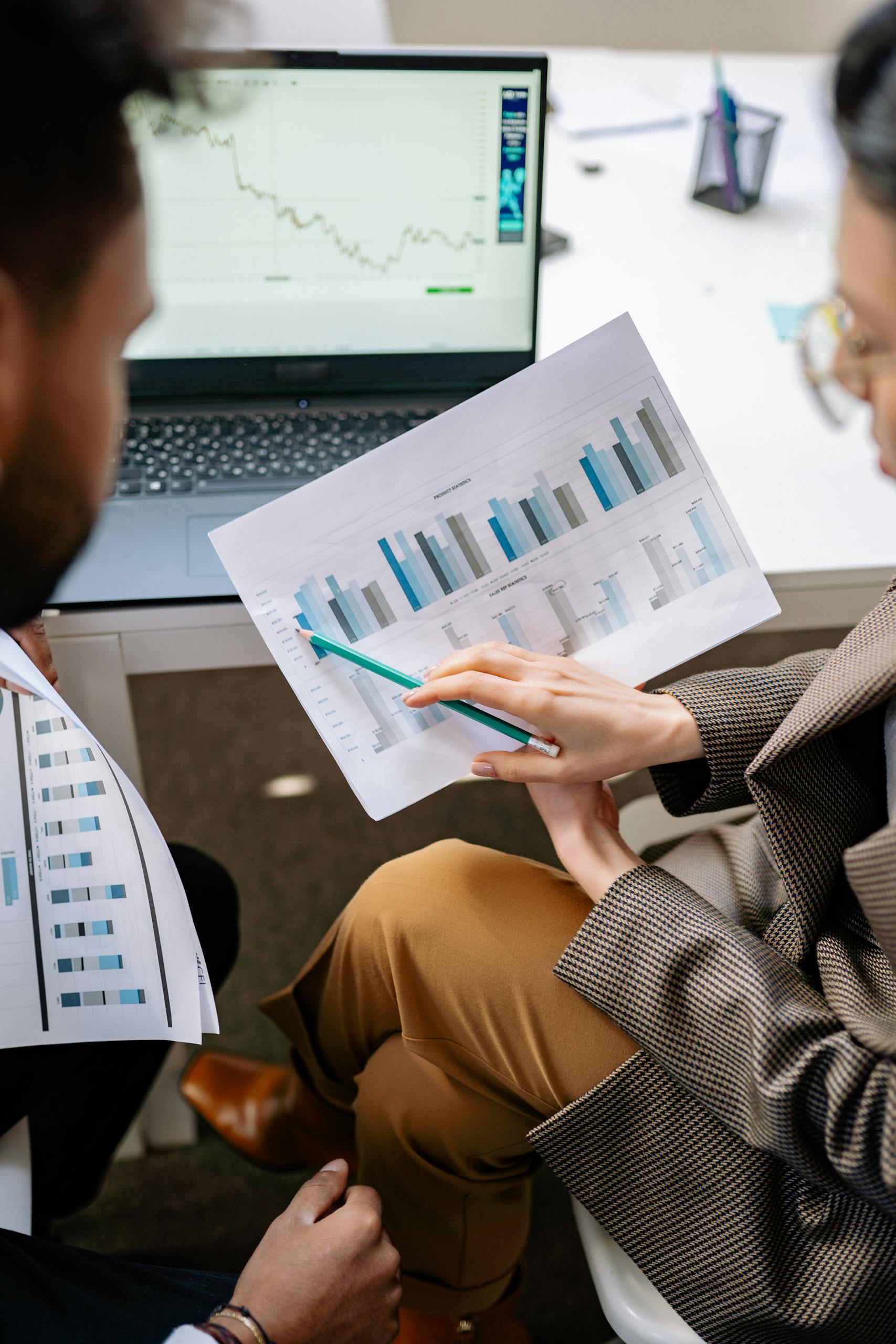 Two adults discuss financial data with charts and laptop in office setting.